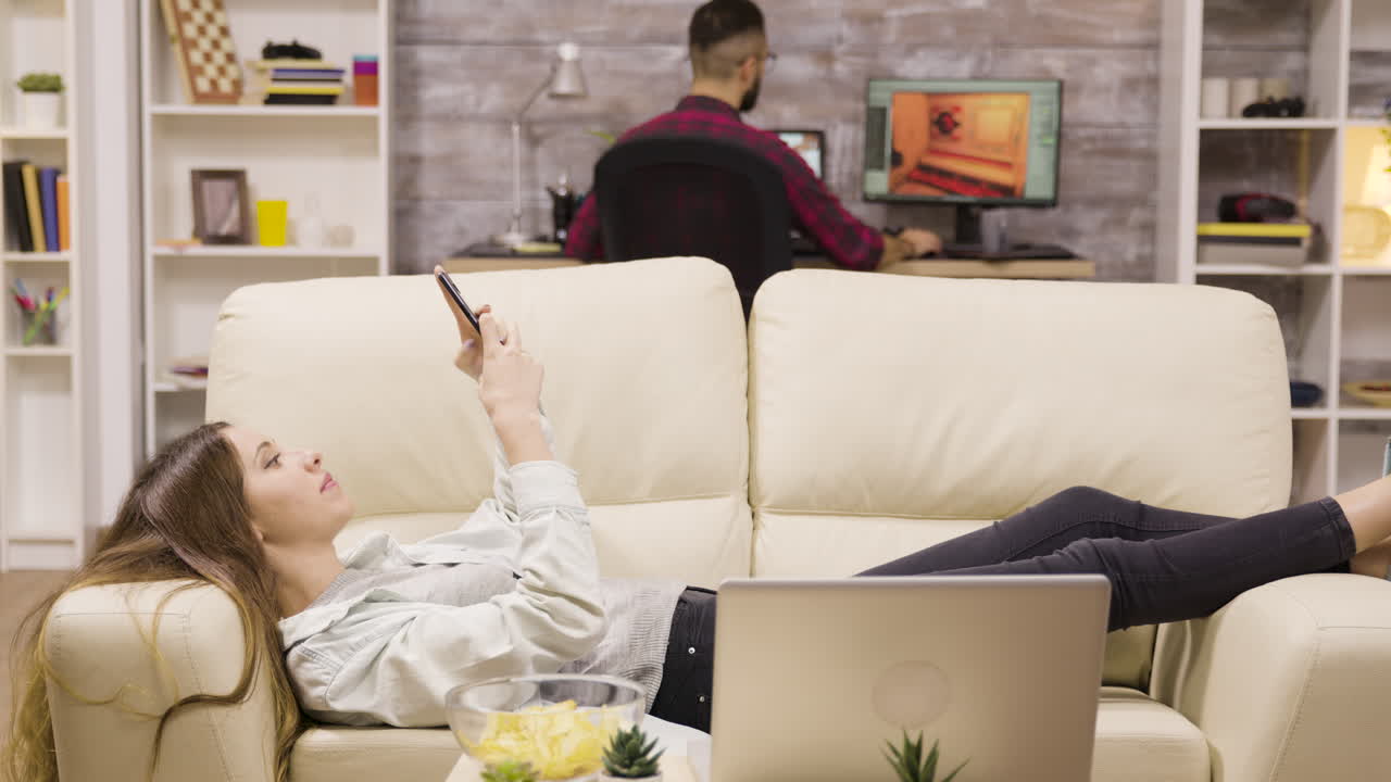 Woman relaxing on couch with smartphone and laptop, man working at computer