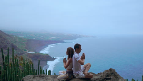 positioned on a mountain's rock, a man and woman sit back to back, finding harmony through meditation and yoga, against the ocean's expanse