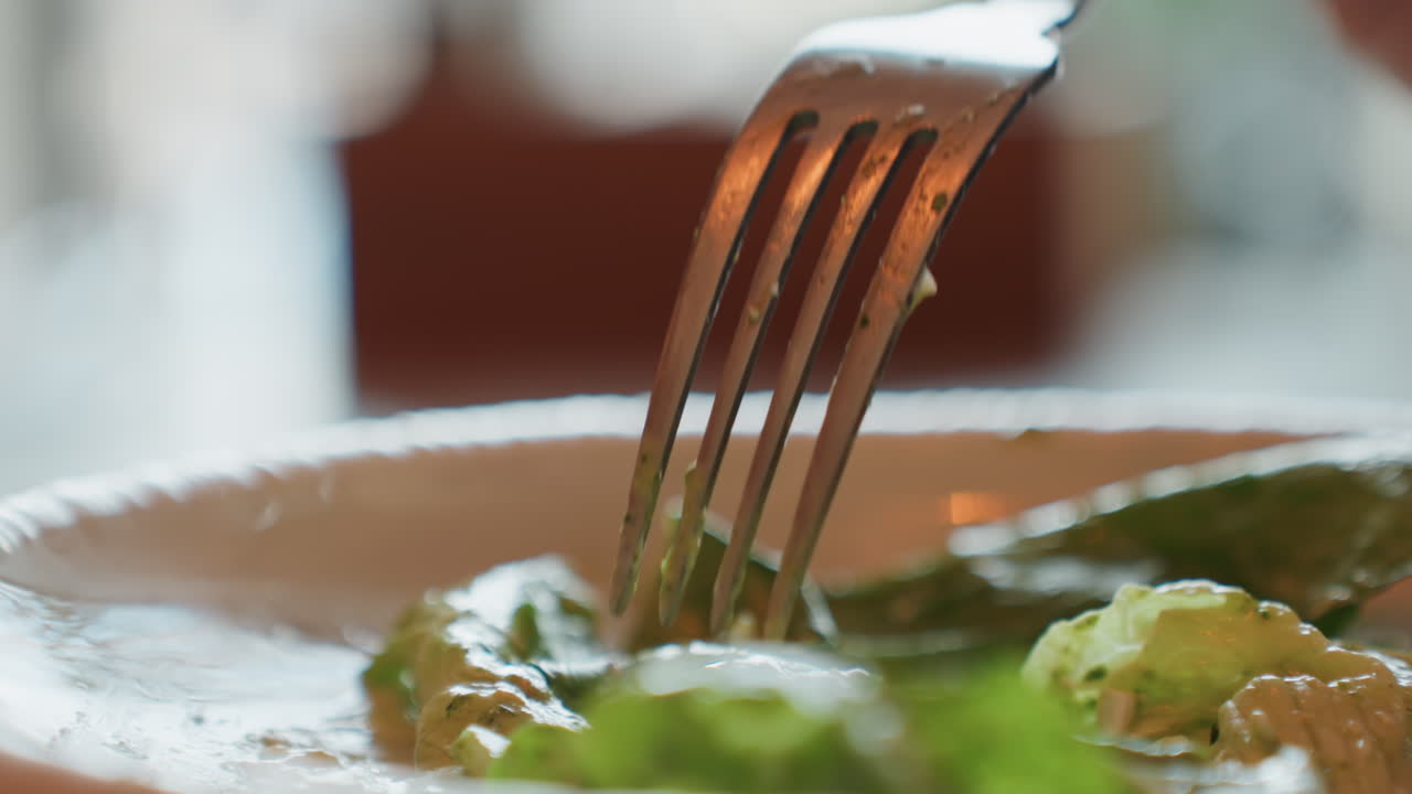 Primo piano di una forchetta che trafigge lattuga romana su un piatto bianco, in un ambiente accogliente di ristorante, i rebbi della forchetta che premono foglie di basilico croccanti con condimento cremoso, sfondo bokeh caldo, ripresa cinematografica al rallentatore