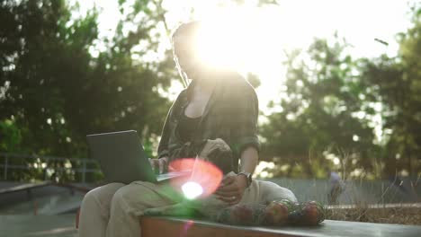 A-woman-with-a-laptop-sits-on-parapet-in-a-skate-park-with-her-dog-pug.-Lens-flares-on-background