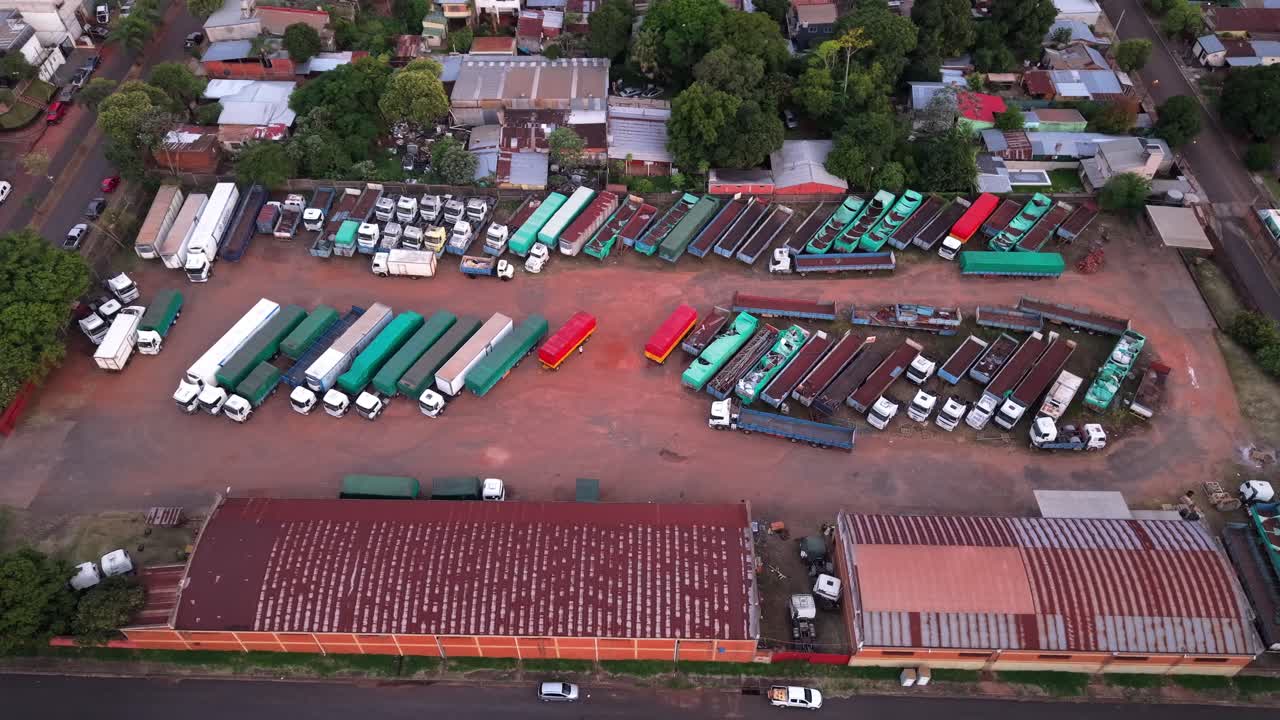 Large red-earthed industrial storage yard packed with semi-trucks, colorful trailers, and corrugated metal buildings, hinting at logistics and transport decay, drone pan and reveal