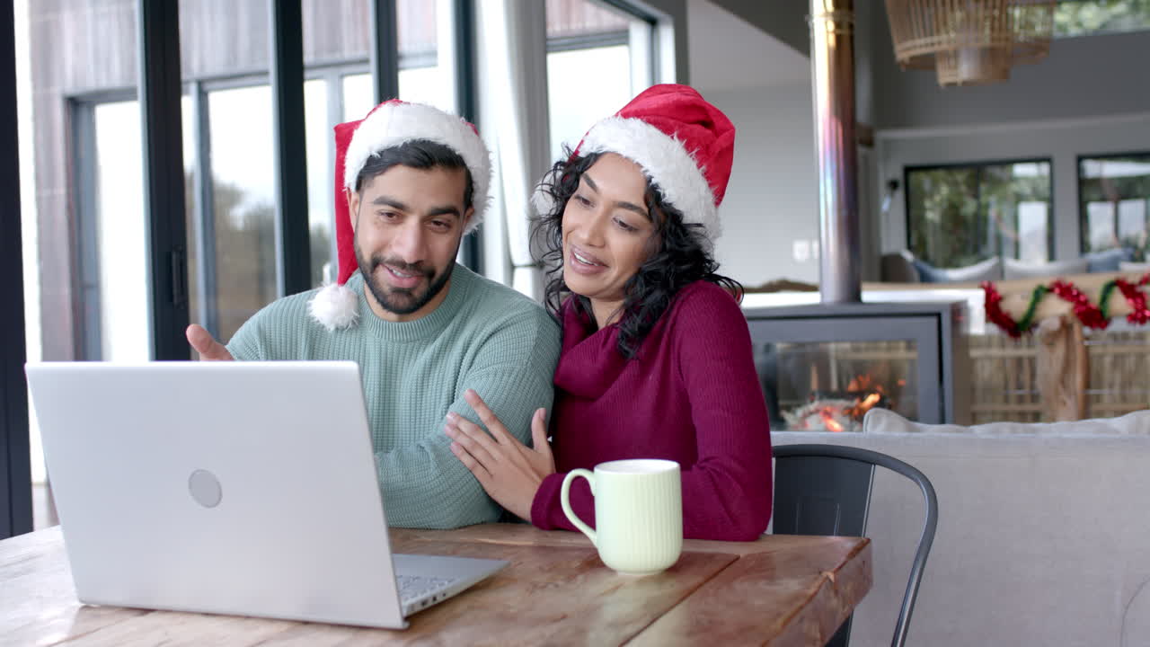 una feliz pareja biracial con sombreros de papá noel usando una computadora portátil para una llamada de video en casa, en cámara lenta