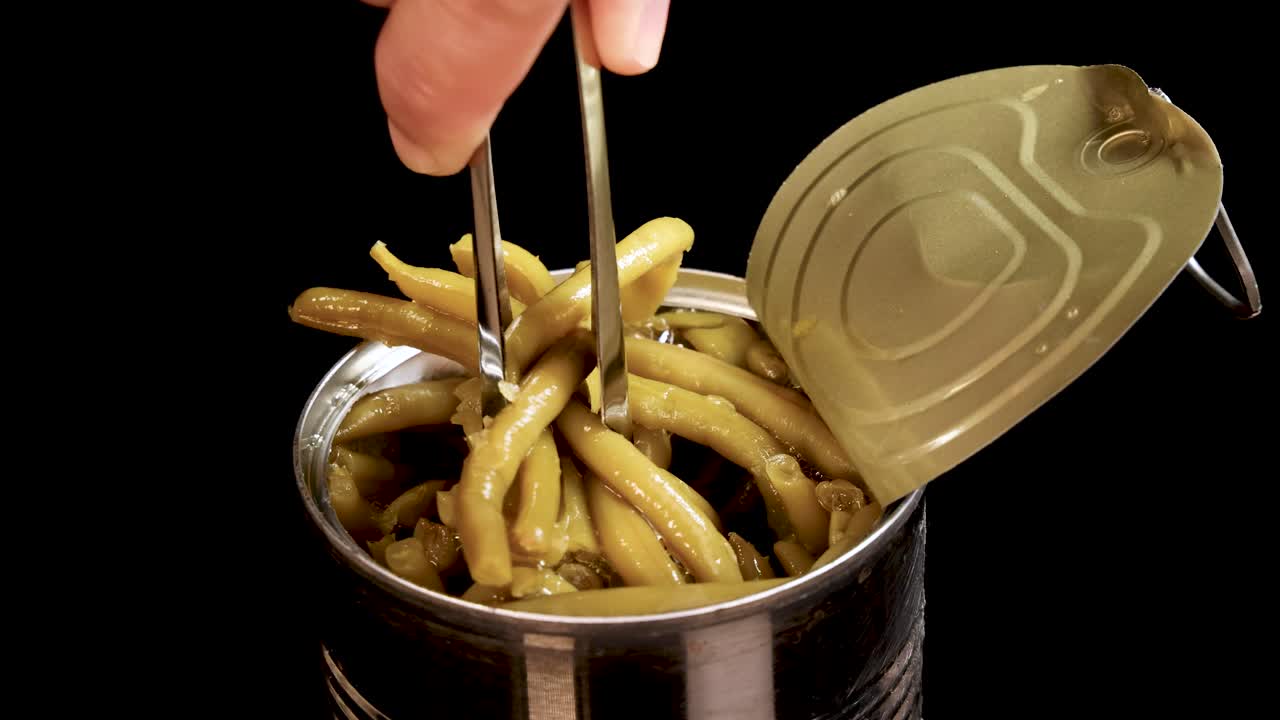 Hand uses metal tongs to lift cooked haricot green beans from an open canned food container against a black background with bright, even lighting