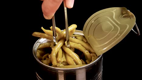 hand uses metal tongs to lift cooked haricot green beans from an open canned food container against a black background with bright, even lighting