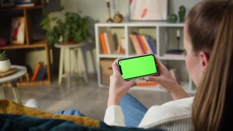 woman using smartphone with green screen. unrecognizable female person holding mobile phone with chroma key, sitting on sofa in living room. watching video or playing game. spare time at home