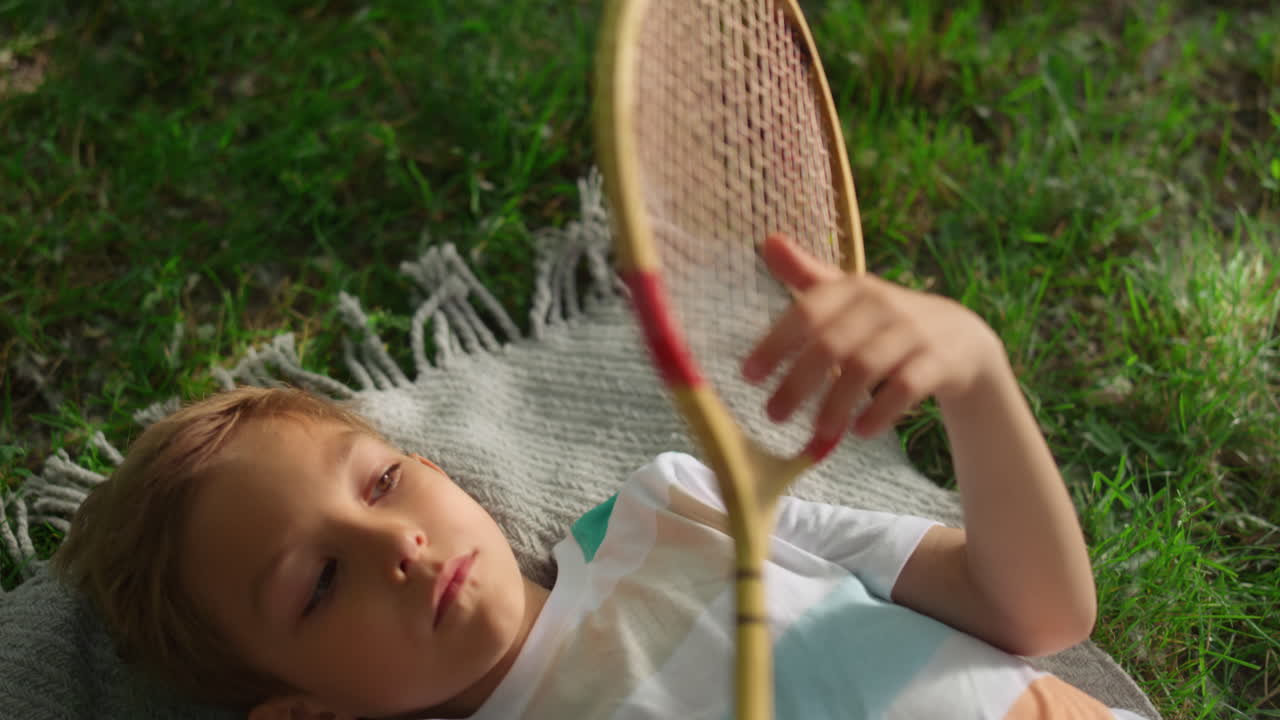 Closeup upset boy examining wooden badminton racket lying green grass alone