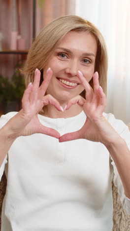 adult woman making heart shape with hands at home smiling gently expressing kindness affection