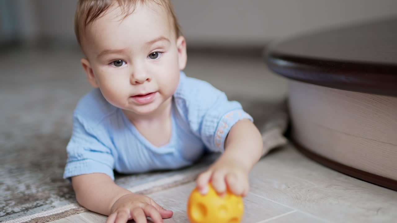 Nice cute baby boy lies on the floor by the stairs playing with a toy. Adorable child hitting ball by the floor. Close up. Blurred backdrop.