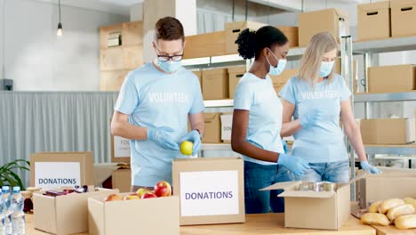 multiethnic group of volunteers in facial mask packing boxes with food and clothes in charity warehouse