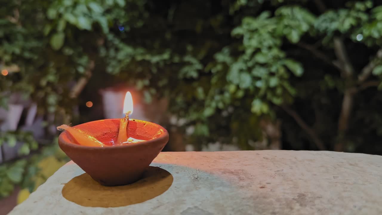 A single Diwali diya glows warmly in a clay lamp as its flame flickers against a softly blurred backdrop of trees, capturing a peaceful festive night atmosphere outdoors