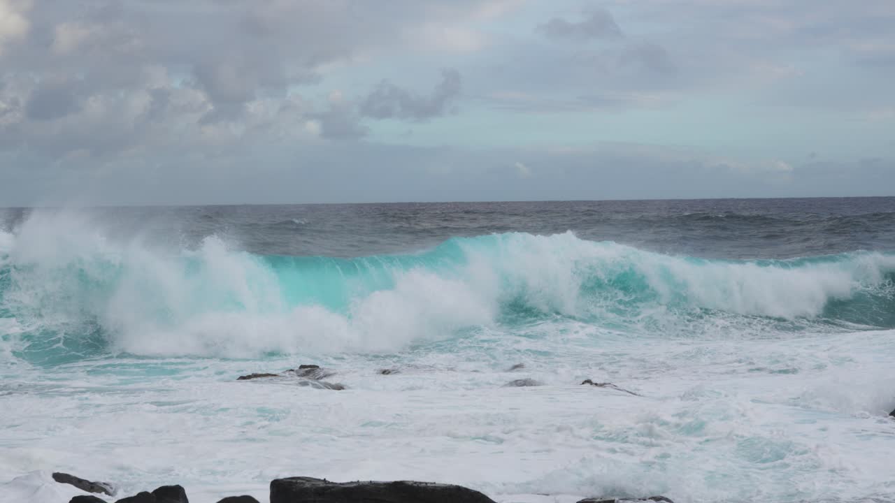 A deep teal set rears and pitches toward shore, detonating into whitewater across nearshore rocks while foamy surge floods the frame beneath a mottled, cloud-swept sky and steady onshore wind