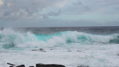 a deep teal set rears and pitches toward shore, detonating into whitewater across nearshore rocks while foamy surge floods the frame beneath a mottled, cloud-swept sky and steady onshore wind