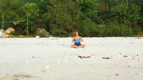 Mujer-Joven-Concentrándose-En-Ejercicios-De-Yoga-Sentada-En-La-Arena-Blanca-De-Una-Playa-Exótica-Frente-Al-Mar-En-Un-Día-Ventoso