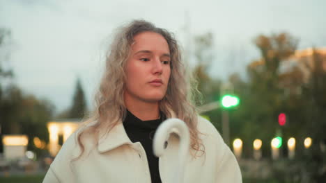 sober lady walking alone looking unhappy holding white umbrella against blurred background filled with bokeh lights trees urban structures during quiet evening stroll under soft moody sky atmosphere