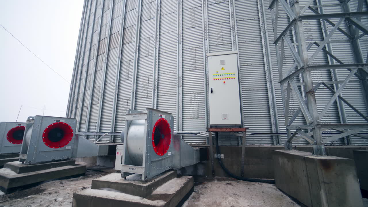 Big ventilators placed outside a huge cylinder of grain elevator. Movement from metal support near grain elevator to the back of one ventilator.