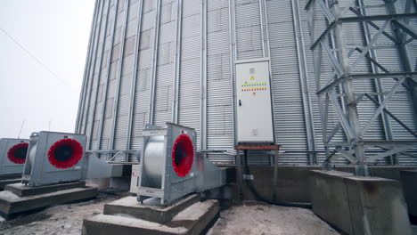 big ventilators placed outside a huge cylinder of grain elevator. movement from metal support near grain elevator to the back of one ventilator.