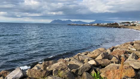 Rocky-shore-with-calm-sea-against-mountain-backdrop-in-Iceland,-under-cloudy-sky