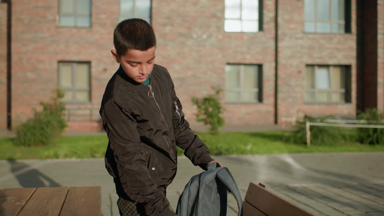 Boy approaches bench with backpack as paper slips from pocket to ground, preparing to drop bag on wooden seat under sunlight with brick building in background and greenery around
