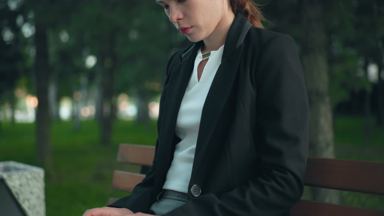 Remote worker seated in calm park on wooden bench focused on laptop with lush greenery and trees in background creating peaceful natural workspace