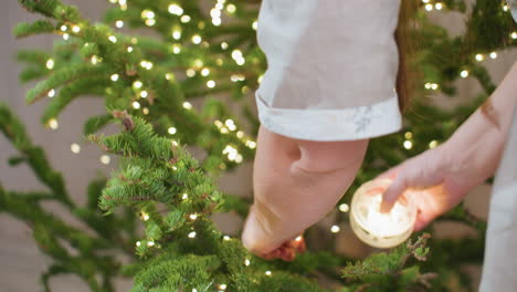 hand view of woman carefully hanging glowing light ornament on christmas tree in cozy festive room filled with soft warm illumination, capturing detailed preparation and joyful holiday spirit indoors