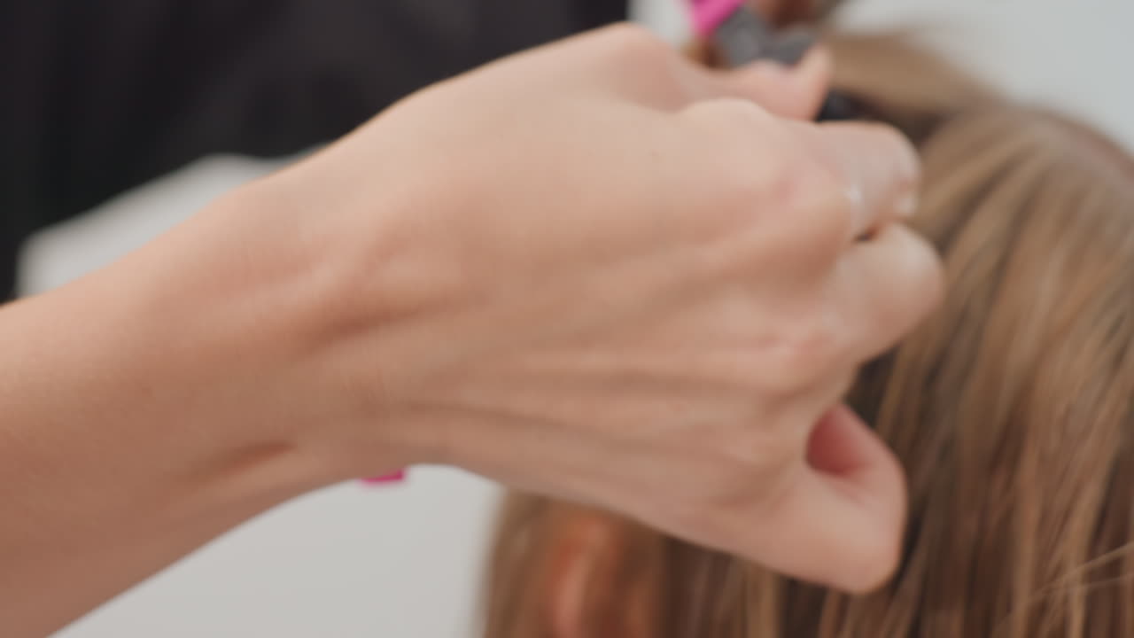Primer plano de unas manos separando mechones de pelo para trenzar; la estilista sujeta pequeñas secciones con clips y prepara microtrenzas y extensiones a lo largo del cuero cabelludo, aplicando una tensión precisa con una técnica manual. Se ven clips y herramientas rosas. Ambiente tranquilo.