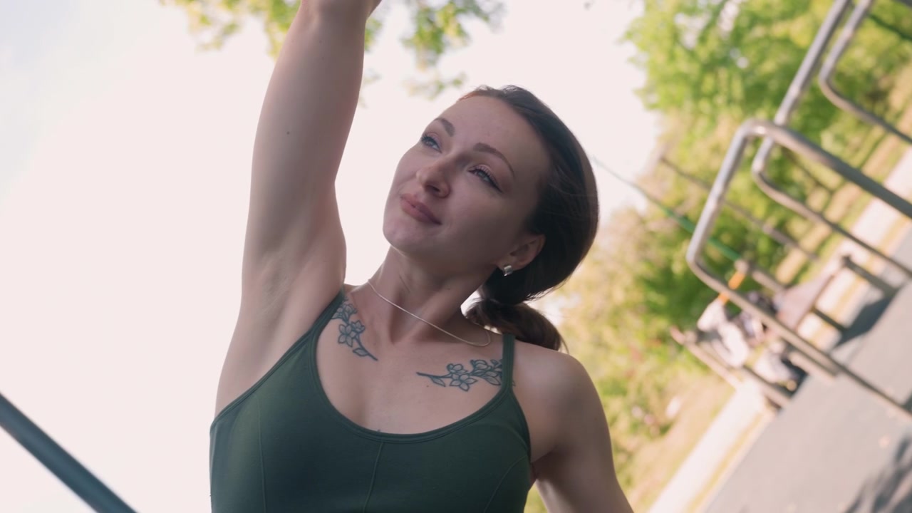 Close-up de uma mulher se alongando ao ar livre à luz do dia, com tatuagens visíveis nos ombros. A mulher veste uma regata verde e realiza alongamentos de braço contra um fundo de parque urbano com elementos de metal.