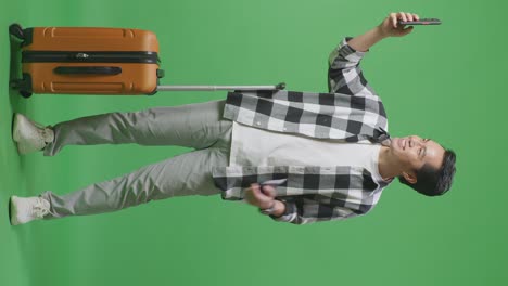 full body of asian male traveler with luggage and passport smiling and taking photo on smartphone while standing in the green screen background studio