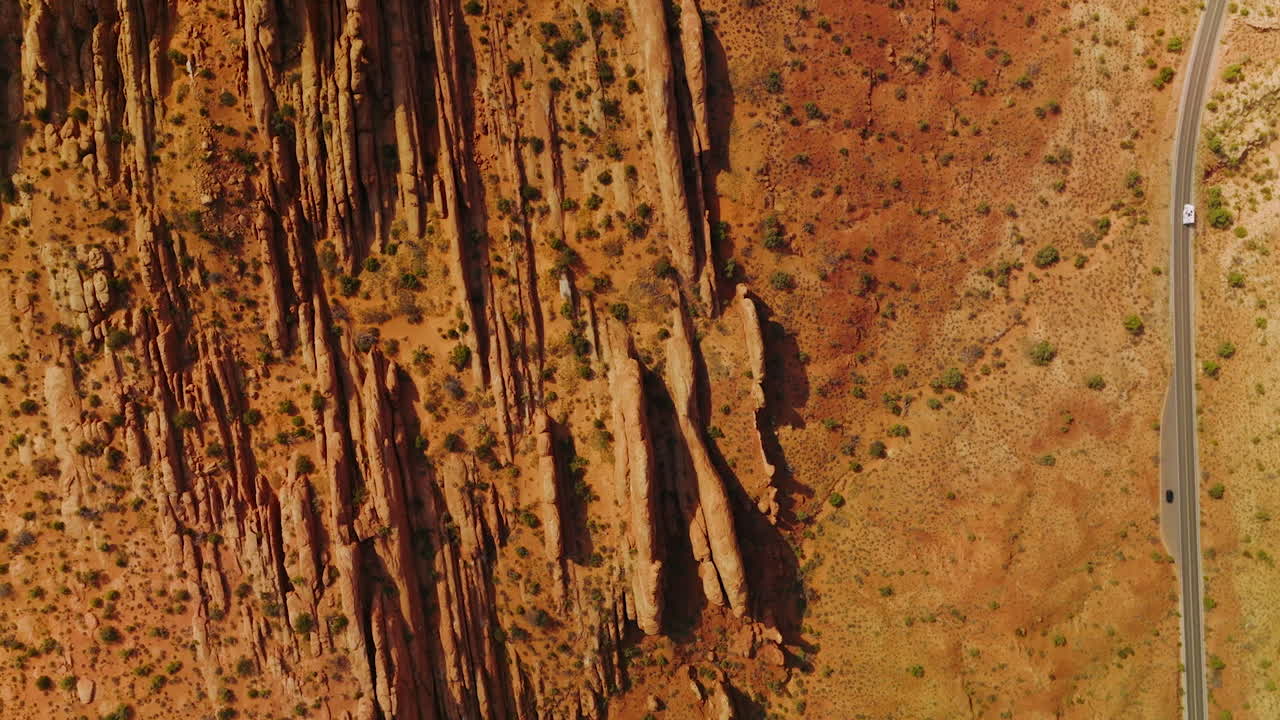 Car going by the road parallel to the oddly-shaped mountains. Dry orange landscape of desert in Utah, USA from aerial view.
