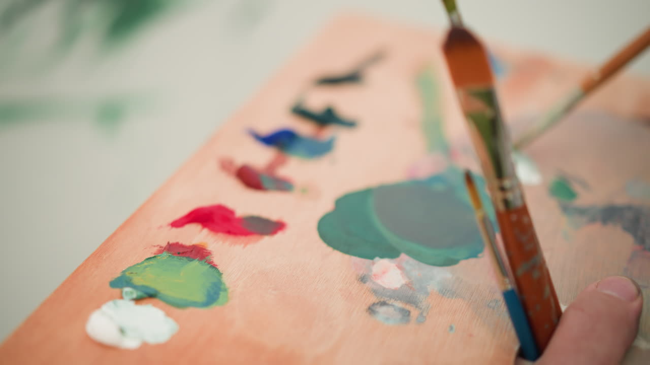 An artist s hand is seen holding a paint palette while delicately mixing paint colors with a brush. The close-up shot captures the careful blending of hues on the wooden palette