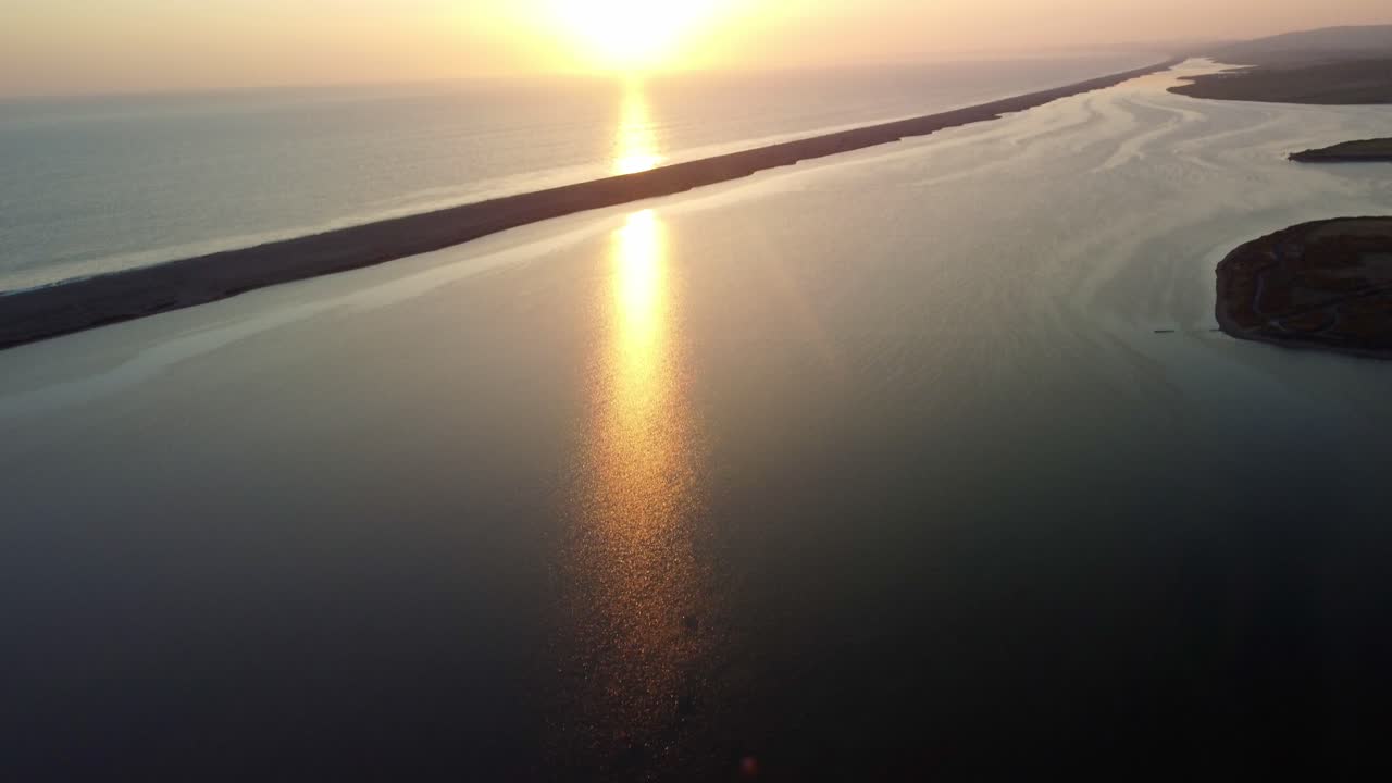 Aerial footage pans up slowly to show a bright, clear sun setting over the sea off Weymouth East Fleet, in Dorset. The sun. reflects across calm water towards the camera. Beautiful yellow and oranges.