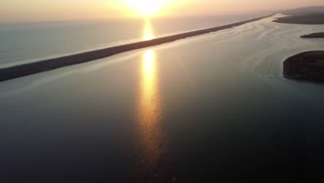 aerial footage pans up slowly to show a bright, clear sun setting over the sea off weymouth east fleet, in dorset. the sun. reflects across calm water towards the camera. beautiful yellow and oranges.