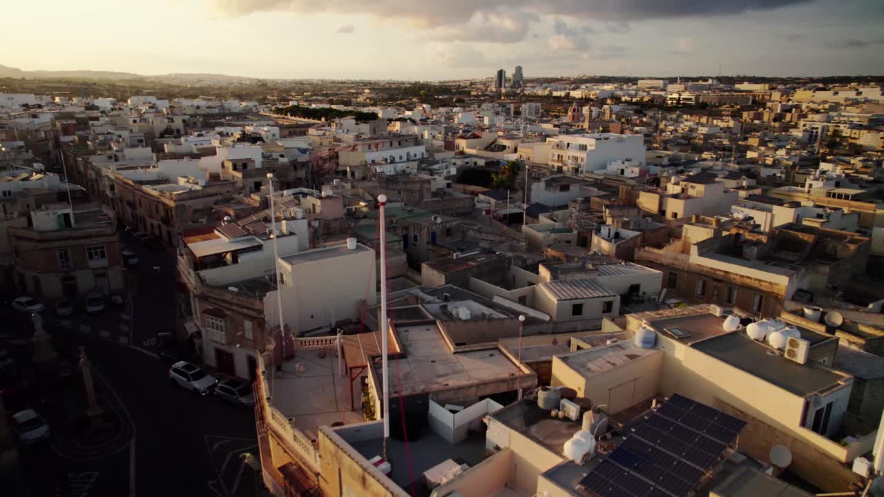 Drone arcs and advances over Qormi, Malta, showing rooftops with solar panels, historic stone buildings, and a church tower glowing in golden sunset light