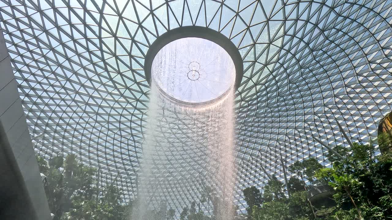 Camera tilts down revealing dramatic indoor waterfall, lush greenery, and geometric glass dome ceiling