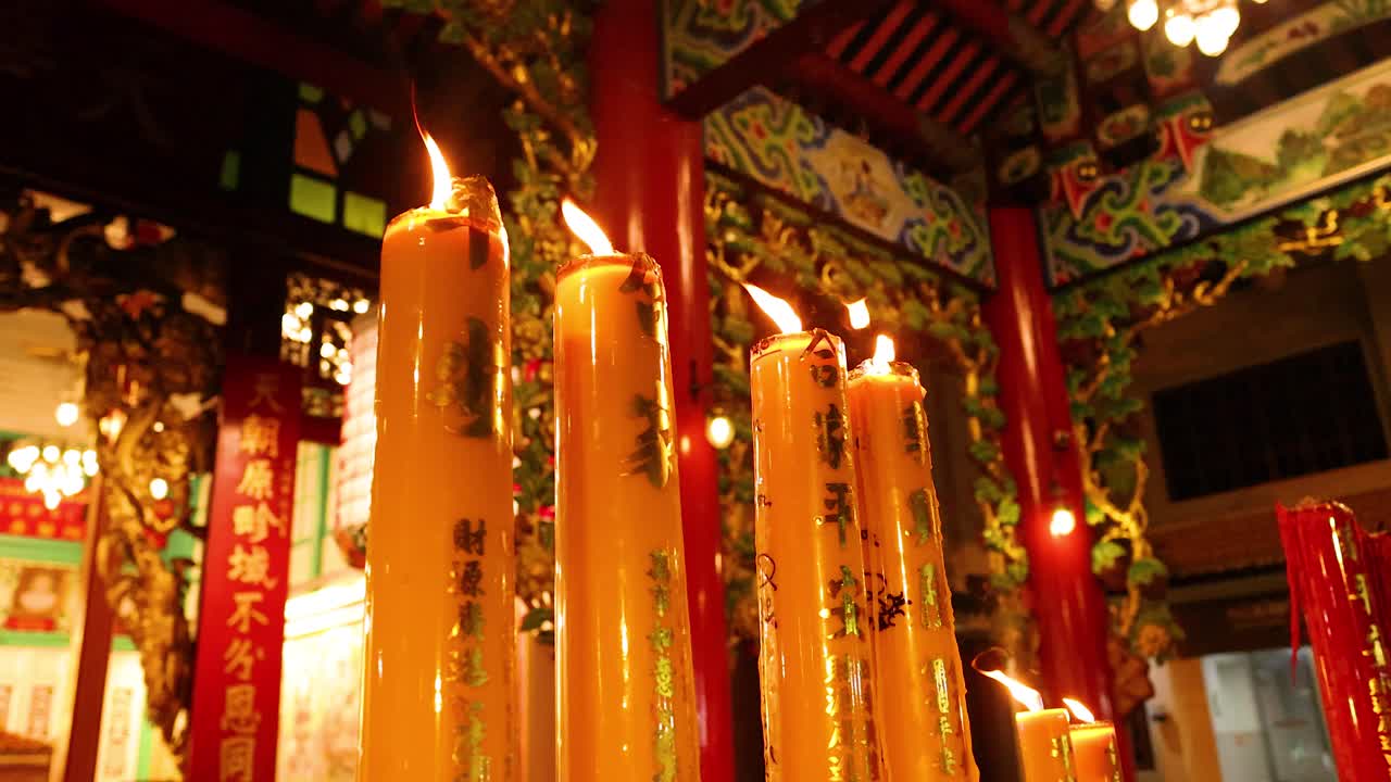 Tall candles burning in richly decorated Asian temple, warm ambient lighting, slight camera movement