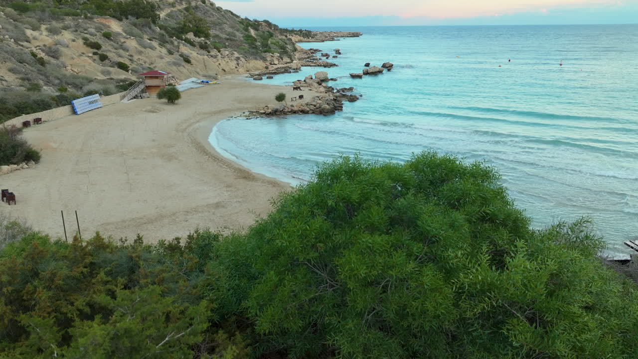 A tranquil scene of a secluded beach as viewed from a vantage point behind a wooden bench and lush greenery, with a glimpse of turquoise waters and a sandy shore nestled between rocky hills, Cyprus