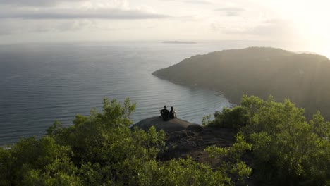 hermoso dron cinematográfico que establece una toma acercándose a una pareja sentada en un punto máximo de senderismo de una montaña de bosque tropical que revela un hermoso paisaje de playa oceánica en el fondo