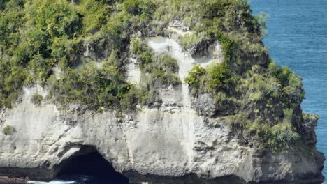 elevándose frente a un gran pináculo de roca marina con cueva, revelando un barco de pesca distante, nusa penida, antena