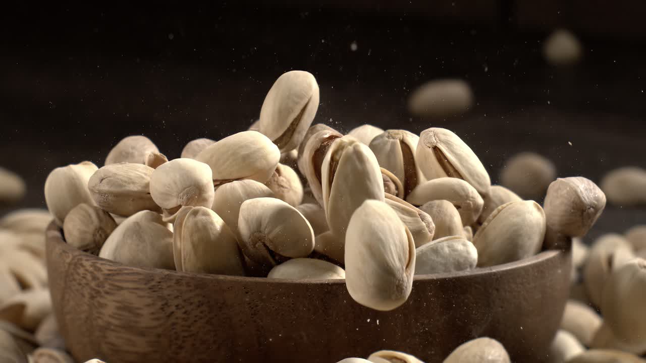 Ciotola di legno piena di pistacchi su tavolo di legno scuro, gustosa frutta secca salata sparsa intorno, concetto di snack salutare e cibo naturale. Scatto con telecamera super slow motion a 1600 fps