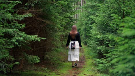 Shot-of-a-woman-with-her-back-is-walking-forward-on-a-narrow-path-between-green-bushes-and-trees-wearing-a-white-skirt-and-a-black-blouse-at-daytime