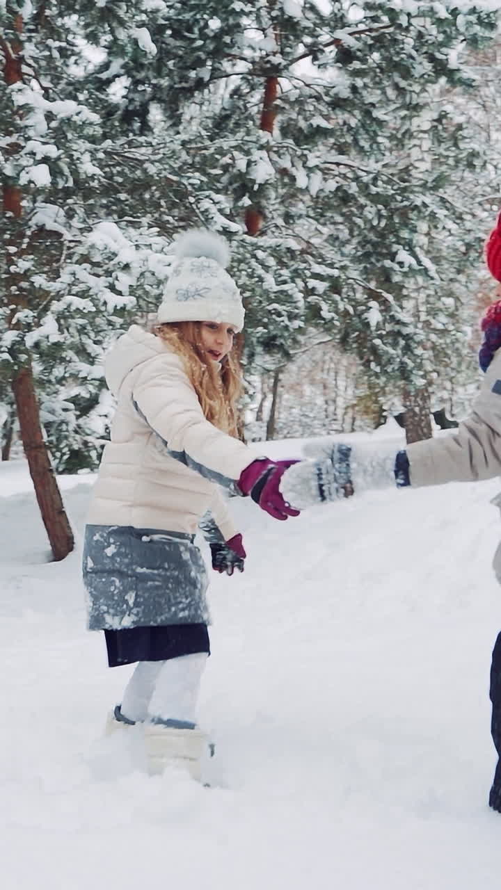 Children are holding each other's hands in the yard and running up in a snowy hill together on the background of trees in the winter. Slow motion Vertical video