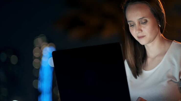 Giovane e bella ragazza designer che lavora da remoto su un laptop per strada in estate, sotto le palme di notte nel parco cittadino. Donna europea negli Emirati Arabi Uniti di notte a Dubai