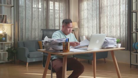 hombre gordo asiático con computadora portátil bebiendo agua mientras trabaja duro con documentos en la oficina
