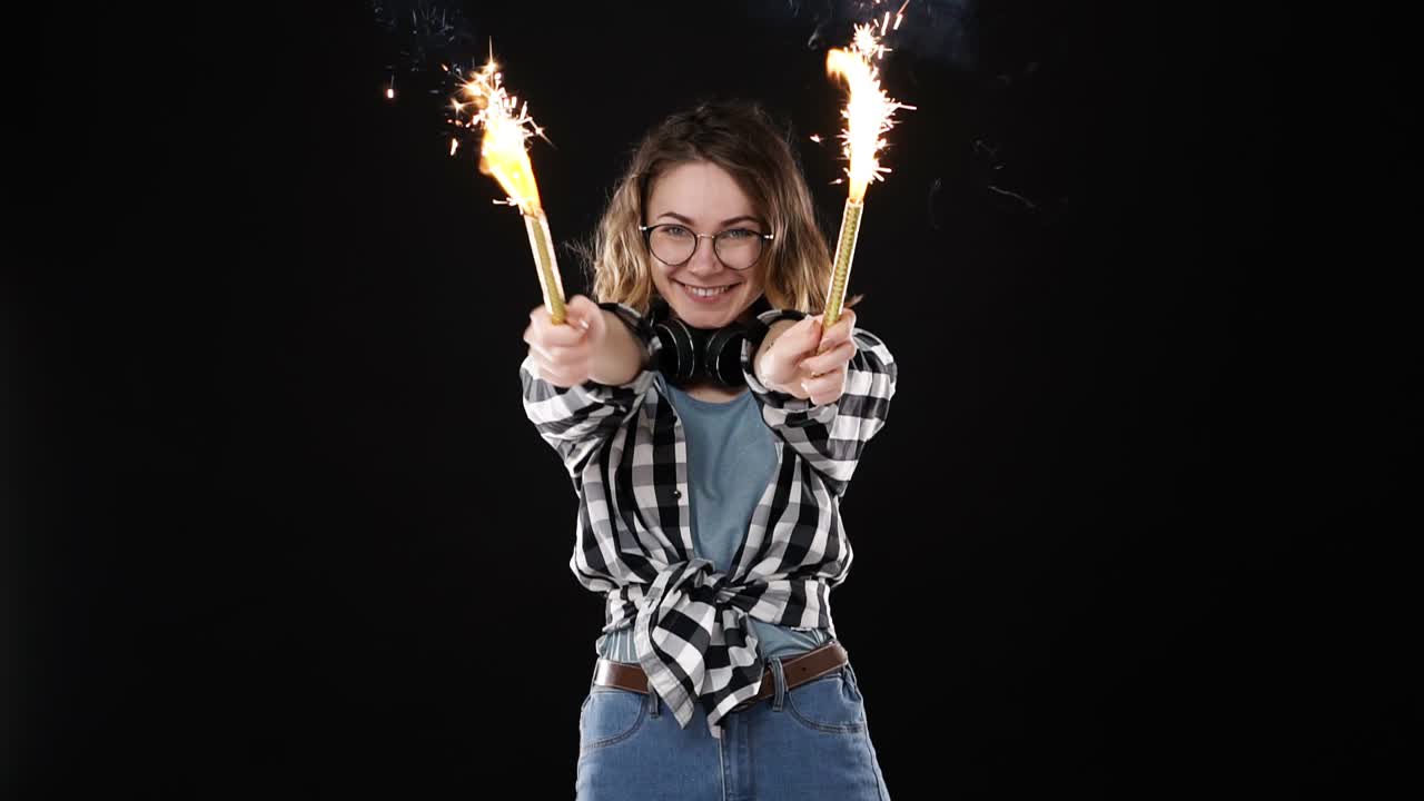 European hipster girl in black and white plaid shirt, headphones on neck celebrating with sparklers and bengal fire. Happy young woman enjoying and having fun against black background. Slow motion