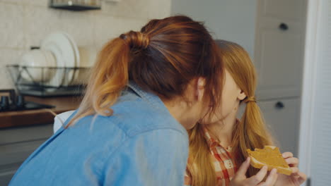 close up of the mother and daughter rubbing noses while a girl eating bread with peanut butter at the kitchen table. portrait shot. indoors