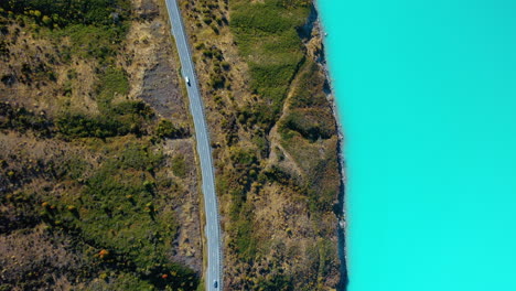 Vista-Aérea-De-Arriba-Hacia-Abajo-De-Un-Automóvil-Conduciendo-Por-Una-Pintoresca-Carretera-De-Montaña-Junto-A-Un-Lago-Glacial-Turquesa