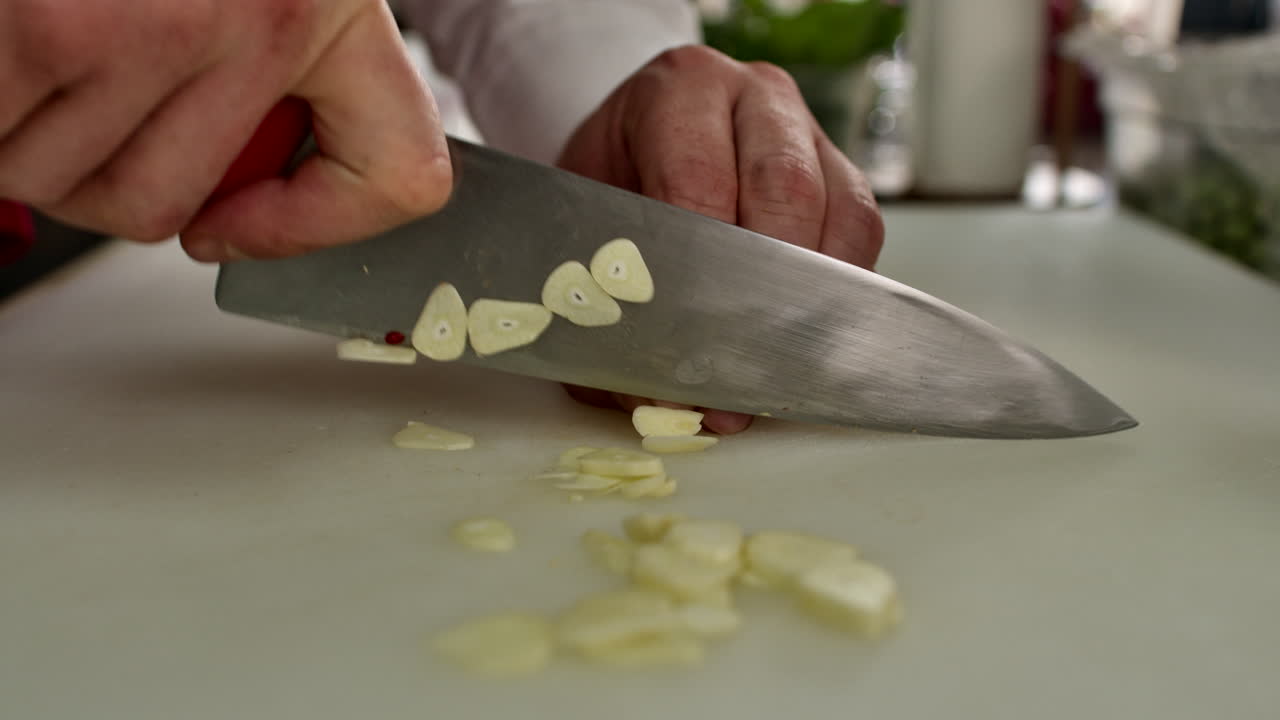 Cinematic close-up shot of a chef's hands finely chopping fresh garlic cloves on a white board. Professional culinary preparation for a tomato sauce recipe