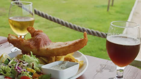 a glass of blonde beer is poured next to a plate of fish, chips, salad, and brown ale on a wooden table in bright natural light