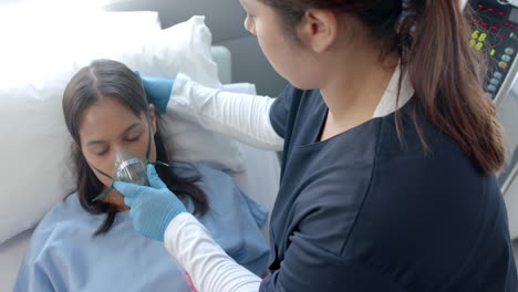 Diverse-female-doctor-putting-oxygen-mask-on-female-patient-in-hospital-bed,-slow-motion