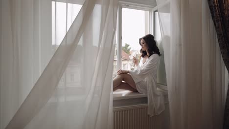 a woman in a white robe sits by a window with a glass of wine, looking out at the city.