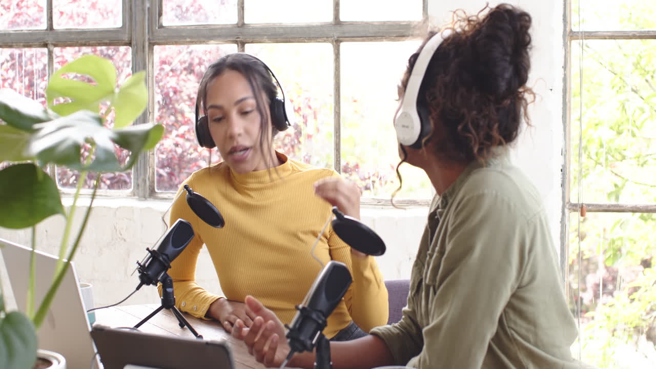 Two multiracial female colleagues recording podcast at home, engaging in lively conversation